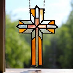 a stained glass cross hanging on a window sill with trees in the back ground