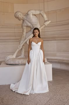 a woman standing in front of a statue wearing a white dress with a bow around her waist