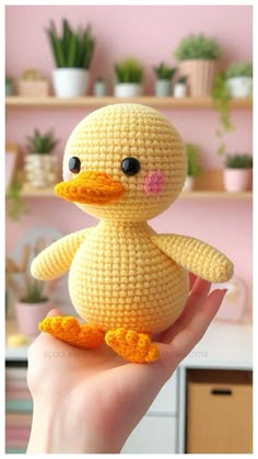 a small crocheted yellow duck sitting on someone's hand in front of a pink wall