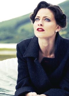 a woman sitting on top of a bed wearing a black jacket and red lipstick with mountains in the background