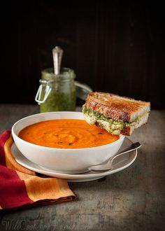 a white bowl filled with soup and a sandwich on top of it next to a jar of pesto