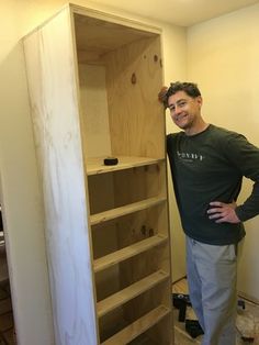 a man standing next to a tall wooden shelf in a room with unfinished walls and flooring