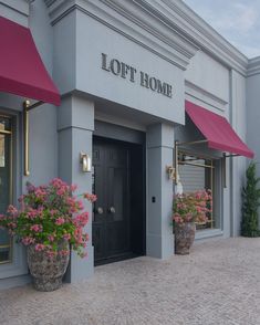 two large planters with pink flowers are in front of a gray building that has red awnings