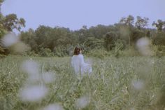 a woman standing in the middle of a field with tall grass and trees behind her