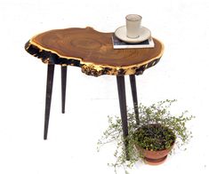 a wooden table with black legs and a white cup on top, sitting next to a potted plant