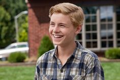 a young man is smiling in front of a brick building with grass and bushes behind him