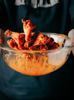 a person holding a glass bowl filled with chicken wings and sauce on top of it