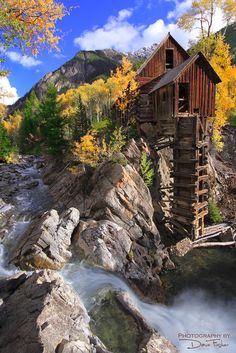 an old wooden water mill on the side of a mountain with fall foliage around it