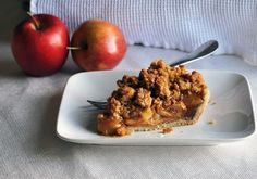 a slice of apple pie on a white plate with a fork and two apples in the background