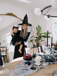 a woman in a witch costume holding a baby on her lap while standing next to a dining room table