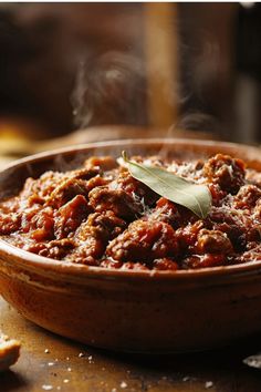 A steaming clay bowl filled with rich, hearty Sunday gravy (Italian meat sauce) garnished with a bay leaf and grated cheese on top. The deep red sauce contains tender chunks of meat, and wisps of steam rise from the rustic earthenware dish, suggesting it's freshly cooked and piping hot. Easy Sunday Gravy Like Nonna Made.