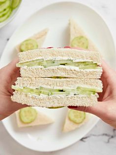 a person holding a sandwich with cucumber slices on it and two bowls of pickles in the background