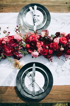 the table is set with silverware and red flowers on it, along with black plates