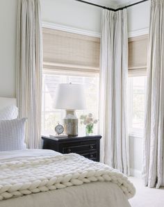 a white bed sitting in a bedroom next to two windows with drapes on them