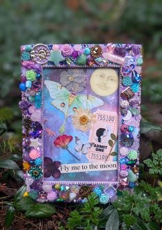 an altered picture frame with flowers, butterflies and a moon in the middle is surrounded by plants