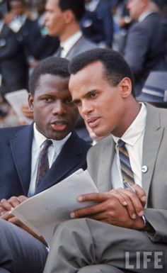 two men in suits sitting next to each other and looking at something on a clipboard