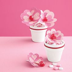 two white flower pots filled with candy hearts on top of a pink table next to each other