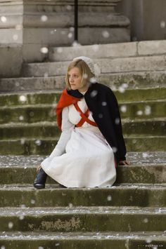 a woman sitting on steps in the snow wearing a white dress and black coat with red trim