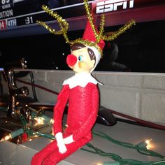 an elf doll sitting on top of a table with christmas lights around him and wearing a santa hat