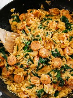 a skillet filled with pasta and sausages on top of a table next to a wooden spoon