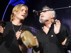 two men standing next to each other on stage with microphones in their hands and one holding a drum