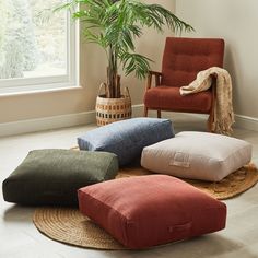 four different colored cushions on the floor in front of a chair and potted plant