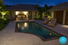 an outdoor swimming pool at night with lounge chairs and thatched roof houses in the background