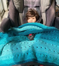 a young boy sitting in a car seat under a blue crocheted blanket on the back of a stroller