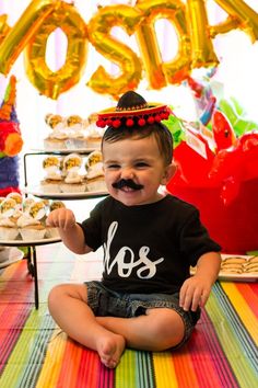 a little boy sitting on the floor in front of some cupcakes and balloons