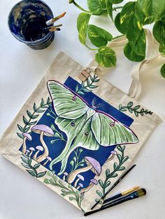 This is a green luna moth hand-painted tote bag with a navy blue background and purple mushrooms. Luna Moth Painting, Moth And Mushroom, Luna Moth Design, Painted Clothes Diy