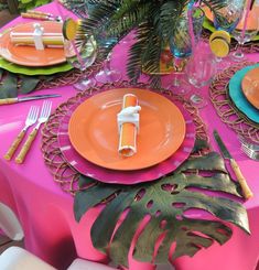 the table is set with pink and orange plates, silverware, and palm leaf napkins