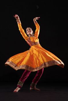 a woman in an orange and gold dress is dancing with her arms spread out to the side