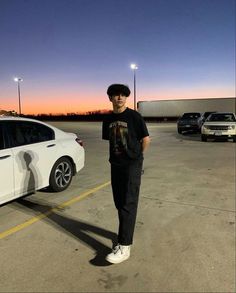 a man standing next to a white car in a parking lot at dusk with the sun setting