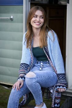 a woman sitting in the doorway of a mobile home wearing ripped jeans and a blue cardigan