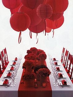 a long table with many red lanterns hanging from it's ceiling and flowers in vases on the tables