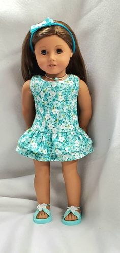 a doll with brown hair wearing a blue dress and sandals, standing on a white sheet