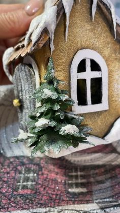 someone is holding a small house with a tree in front of it and snow on the roof