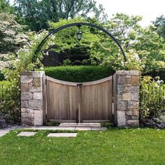 a wooden gate in the middle of a lush green yard with stone walls and pillars