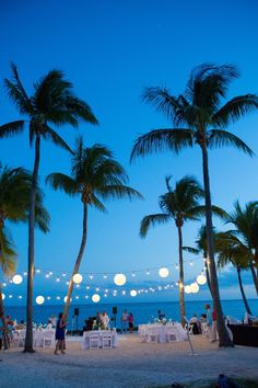 people are sitting at tables on the beach under palm trees with lights strung from them