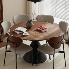 a round wooden table surrounded by chairs and a vase with fruit on it in front of a window