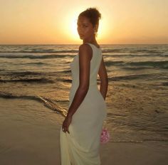 a woman standing on the beach at sunset wearing a white dress and holding a pink flower in her hand