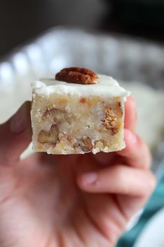 a hand holding a piece of cake with nuts on top in front of a white plate