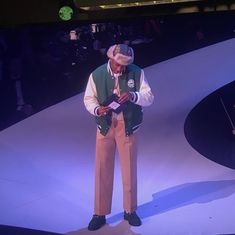 a man standing on top of a stage wearing a green vest and hat with his hands in his pockets