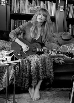 a woman sitting on a couch with a guitar in front of her and bookshelves behind her