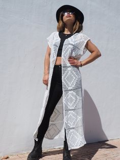 a woman standing in front of a white wall wearing a black hat and long cardigan