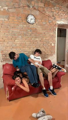 three people sitting on a red couch in front of a brick wall with a clock