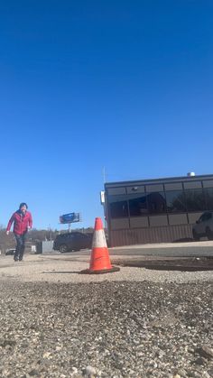 a man in red jacket standing next to orange cone on gravel road with building and blue sky