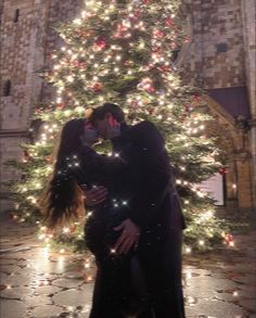 a man and woman kissing in front of a christmas tree with lights all around them