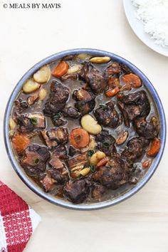 a bowl filled with meat and vegetables on top of a wooden table next to rice