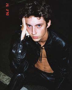 a young man wearing a black leather jacket and holding his hand to his head while sitting on the ground